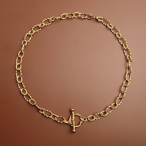 Könnte beinhalten: Eine goldfarbene Halskette mit einem strukturierten Kettendesign und einem Knebelverschluss. Die Kette besteht aus ovalen Gliedern mit einem gedrehten Muster, was ein einzigartiges und auffälliges Accessoire ergibt. Die Halskette wird auf braunem Hintergrund präsentiert.