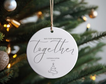 May include: A white ceramic Christmas ornament with the words "Our First Christmas Together" in a script font. Below the text is "Custom & Custom 2023" and a small Christmas tree illustration. The ornament hangs from a twine string.