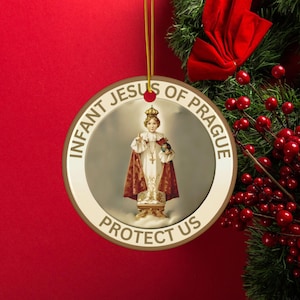 Può includere: Ornamento natalizio rotondo con l'immagine del Bambino Gesù di Praga. L'immagine è circondata da un bordo dorato con la scritta "INFANT JESUS OF PRAGUE PROTECT US". L'ornamento è appeso a un filo d'oro su sfondo rosso.