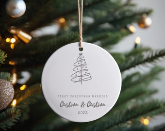 May include: White ceramic Christmas ornament with a minimalist tree design and the text "First Christmas Married" and "Custom & Custom 2023". The ornament hangs from a natural twine string, displayed on a Christmas tree.
