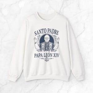 Może przedstawiać: Biała bluza z granatową grafiką. Grafika przedstawia ilustrację mężczyzny z podniesionymi ramionami, otoczonego tekstem "SANTO PADRE PAPA LEÓN XIV". Projekt zawiera elementy dekoracyjne i książkę.