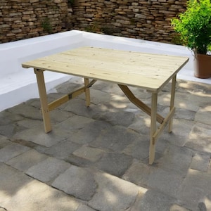 May include: A light-coloured wooden folding table with a rectangular top and angled legs. The table is made of unfinished wood and is set up outdoors on a stone patio. The table is approximately 122 cm long.