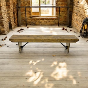 May include: A wooden bench with a rectangular top and angled legs. The bench is made of light brown wood with visible grain and is supported by black metal brackets. The setting is a room with brick walls and a wooden floor.