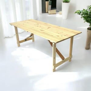 May include: A light wood folding table with a simple design. The table is made of natural wood and has a rectangular top. The table is supported by four legs that fold in for easy storage.
