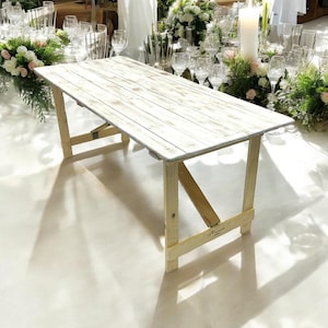 May include: A whitewashed wooden table with a rustic finish. The table has a simple, rectangular design with a wooden frame and legs. The table is ideal for a wedding or other special event.