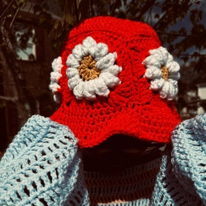 May include: A red crocheted bucket hat with white daisy flowers. The hat is worn over a blue and pink striped crocheted top.