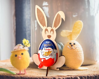 May include: Wooden bunny rabbit Easter egg holder with a red and blue Kinder Joy chocolate egg inside. The bunny is holding the egg in its paws. There is a yellow chick with a pink hat in the foreground.