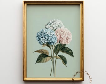 May include: A framed floral print featuring three hydrangea blooms in shades of blue, pink, and white. The artwork is set against a soft green background and framed in a gold-colored frame. The text "THE SEASONED GALLERY" is printed at the top and bottom.