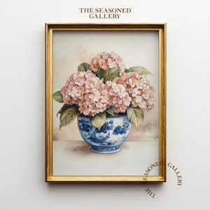 Puede incluir: Una impresión enmarcada de acuarela con un ramo de hortensias rosas en un jarrón de porcelana azul y blanco. El jarrón tiene un estampado floral. El marco es dorado. El texto "THE SEASONED GALLERY" está en la parte superior e inferior derecha.