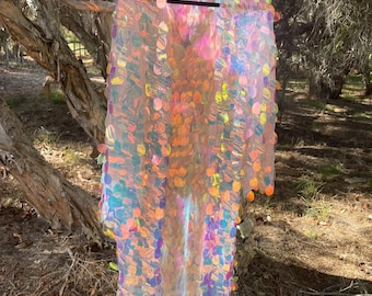 Puede incluir: Una prenda larga y transparente adornada con numerosas lentejuelas circulares iridiscentes de colores brillantes. Cuelga de una percha negra sobre un fondo natural de ramas y follaje. Las lentejuelas crean un efecto brillante.