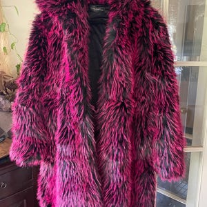May include: A long faux fur coat in a striking combination of hot pink and black. The coat features a hooded design and a plush, textured appearance, perfect for a bold fashion statement. The coat is hanging on a hanger.