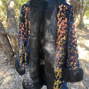 May include: A long black coat with a faux fur collar and trim. The coat is embellished with colourful sequins, creating a shimmering effect. The coat is hanging on a black hanger.