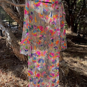 May include: A sheer, long-sleeved, open-front garment with colorful sequin embellishments. The sequins are arranged in floral patterns, featuring shades of pink, orange, blue, and green. The garment hangs on a black hanger against a natural outdoor backdrop.