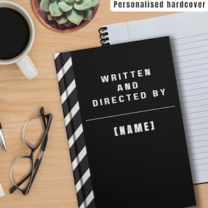 Puede incluir: Cuaderno negro y blanco con una cubierta personalizada de tapa dura. La cubierta tiene un diseño a rayas en blanco y negro y el texto "Written and Directed By [NAME]" en blanco.