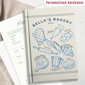 Könnte beinhalten: Ein Rezeptbuch mit beigefarbenem Hardcover und dem Text "BELLA'S BAKERY EST. 2023" in blauer Schrift. Der Umschlag zeigt blaue Illustrationen von Backutensilien und Zutaten, darunter ein Nudelholz, ein Schneebesen, ein Spritzbeutel, ein Messbecher, ein Schneidebrett, ein Brotlaib, ein Croissant und Ofenhandschuhe.