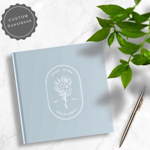 May include: A light blue guest book with a white logo that says "Cozy Home Apartment" with a floral design and the text "Est. 2024".