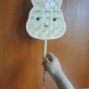 May include: A pink and brown woven bunny-shaped hand fan with blue eyes and a pink flower on top. The fan is attached to a wooden stick with a pink fluffy ball on a string.