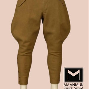 Puede incluir: Pantalones de montar color canela con detalles de botones en la pantorrilla y solapas en los bolsillos. Los pantalones tienen una cintura alta y una forma distintiva y completa en los muslos. El logo "MAANMUK Dress to Succeed" es visible.