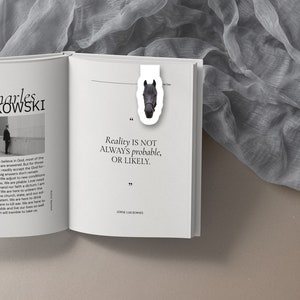 May include: A white bookmark with a black horse head illustration. The bookmark is placed on a page of an open book. The text on the page reads "Reality is not always probable, or likely." and is attributed to Jorge Luis Borges.
