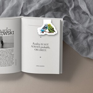May include: A white bookmark with a quote by Jorge Luis Borges: "Reality is not always probable, or likely." The bookmark features a colorful illustration of two parakeets.