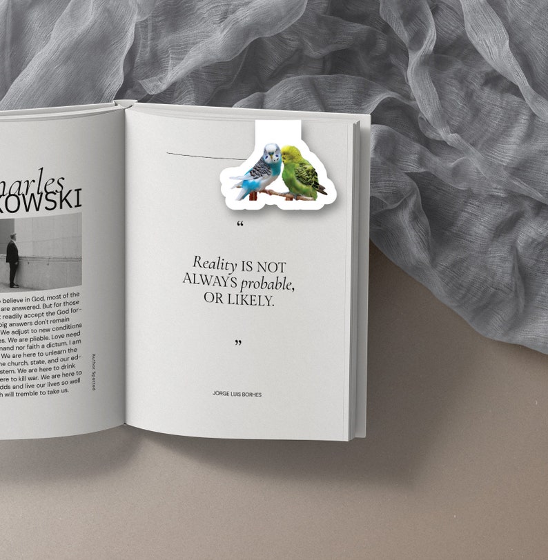 May include: A white bookmark with a quote by Jorge Luis Borges: "Reality is not always probable, or likely." The bookmark features a colorful illustration of two parakeets.