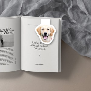 May include: A white bookmark with a golden retriever dog head illustration. The bookmark has the quote "Reality is not always probable, or likely." by Jorge Luis Borges.