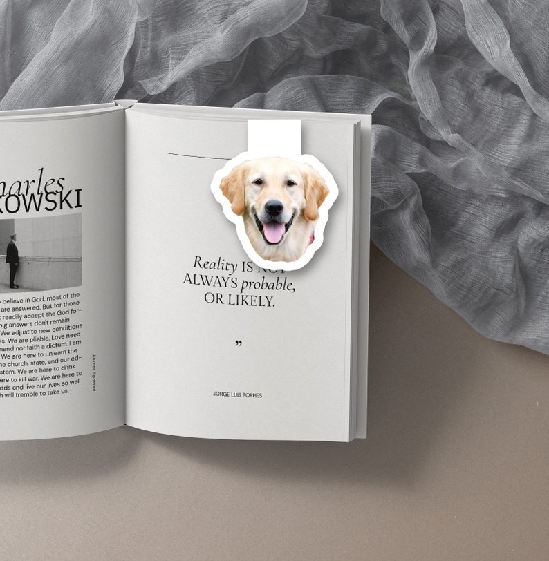 May include: A white bookmark with a golden retriever dog head illustration. The bookmark has the quote "Reality is not always probable, or likely." by Jorge Luis Borges.