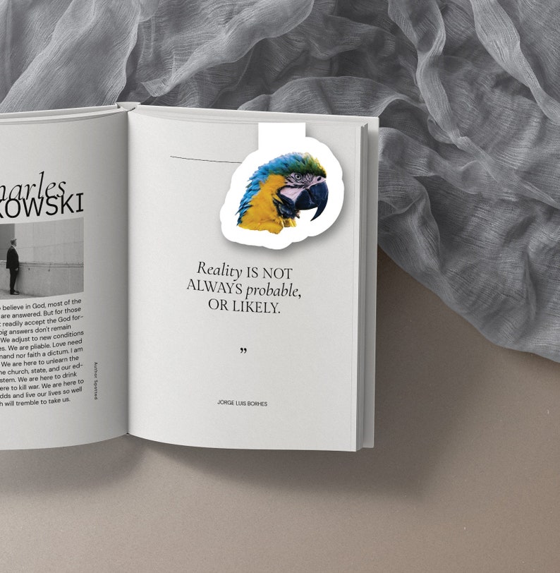 May include: A blue and yellow macaw with a black beak is featured on a white bookmark. The bookmark is placed on an open book with the text "Reality is not always probable, or likely." and "JORGE LUIS BORHES".