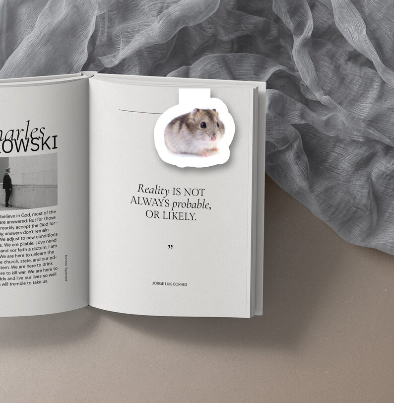 May include: A white bookmark with a quote by Jorge Luis Borges: "Reality is not always probable, or likely."  The bookmark features a small, white hamster with brown markings.
