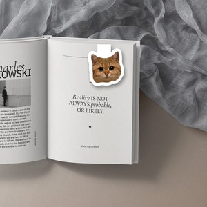 May include: A white bookmark with a picture of a ginger cat on it. The bookmark is on a page of a book with the quote "Reality is not always probable, or likely." by Jorge Luis Borges.