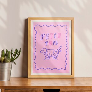 May include: A framed print with a pink background and a white border. The print features a cartoon dog with the text "FETCH THIS" in purple and pink letters.