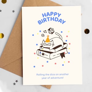 May include: A birthday card with a cartoon cat wearing a party hat, sitting on books, and surrounded by dice and confetti. The card reads "Happy Birthday" and "Rolling the dice on another year of adventure!"