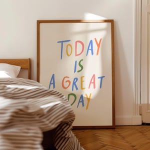 May include: A framed art print with the words "TODAY IS A GREAT DAY" in colorful, handwritten-style lettering. The artwork is set against a cream background and framed in a wooden frame. The print is displayed near a bed with striped bedding.