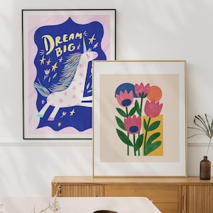 May include: Two framed art prints. One features a unicorn with the text "DREAM BIG" on a blue background. The other print has pink flowers, green leaves, and a yellow rectangle on a beige background. Both are in gold frames.