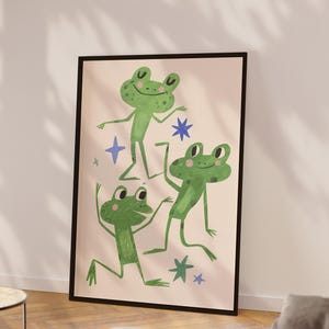 May include: A framed art print featuring three whimsical green frogs in playful poses against a light pink background. The frogs are surrounded by blue and green star shapes. The artwork is framed in a black border.