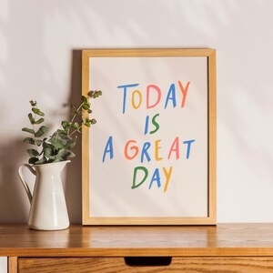 May include: A framed print with the words "TODAY IS A GREAT DAY" in colorful, handwritten-style letters. A white pitcher with green foliage sits to the left of the frame on a wooden surface.