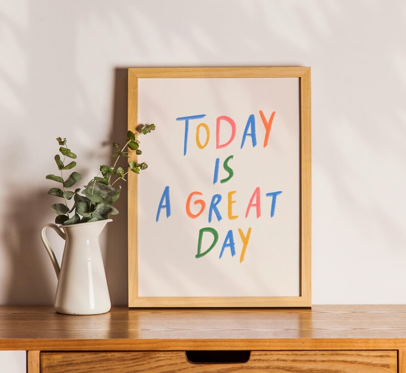 May include: A framed print with the words "TODAY IS A GREAT DAY" in colorful, handwritten-style letters. A white pitcher with green foliage sits to the left of the frame on a wooden surface.
