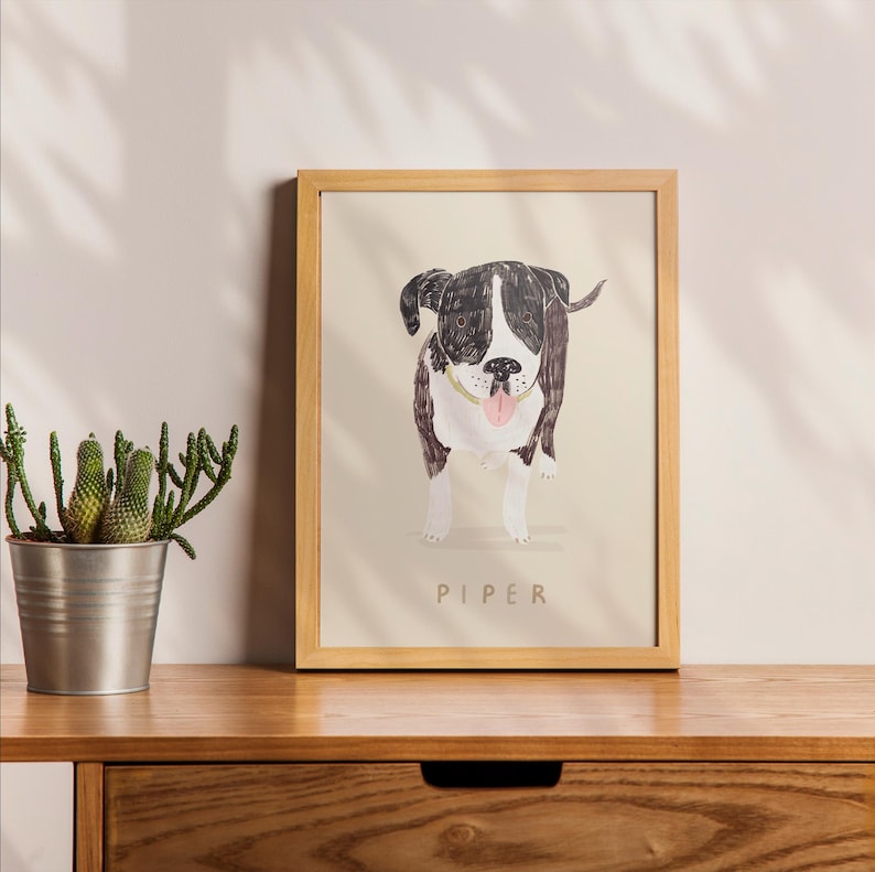 May include: A framed illustration of a black and white dog with a pink tongue sticking out. The dog is looking at the camera and the frame is made of light wood. The name "PIPER" is printed below the dog.