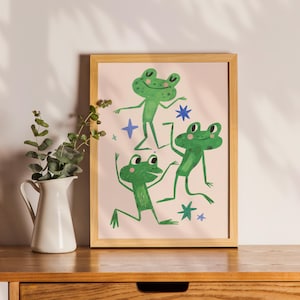 May include: A framed print featuring three green cartoon frogs in playful poses against a light pink background. The frogs are depicted with smiling faces and are surrounded by blue star and cross shapes. The artwork is displayed on a wooden surface.