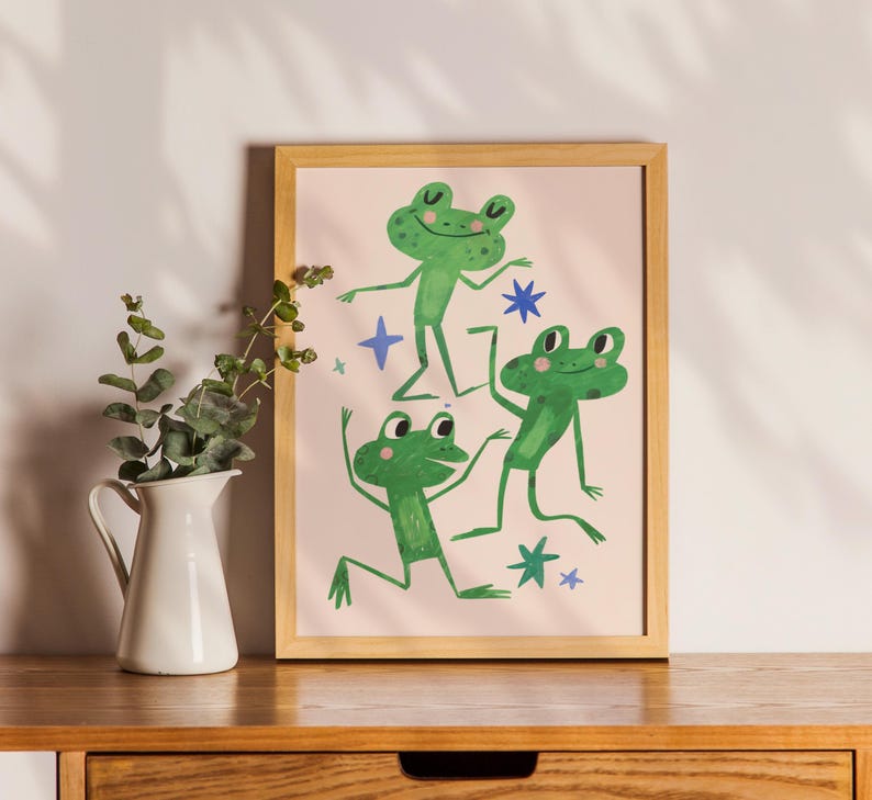 May include: A framed print featuring three green cartoon frogs in playful poses against a light pink background. The frogs are depicted with smiling faces and are surrounded by blue star and cross shapes. The artwork is displayed on a wooden surface.