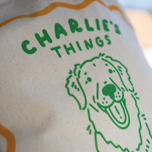 May include: A cream-colored canvas tote bag featuring a green screen-printed graphic of a smiling dog and the text CHARLIE'S THINGS in a bold, rounded font. An orange wavy line border frames the upper portion of the design on the fabric.