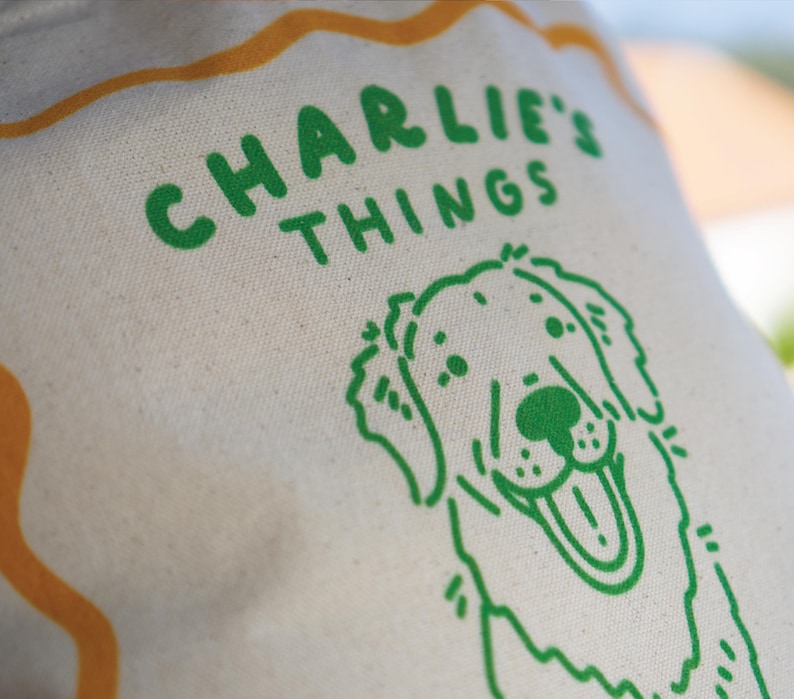May include: A cream-colored canvas tote bag featuring a green screen-printed graphic of a smiling dog and the text CHARLIE'S THINGS in a bold, rounded font. An orange wavy line border frames the upper portion of the design on the fabric.