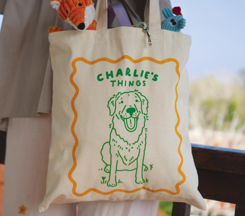 May include: A cream-colored canvas tote bag featuring a green line-art illustration of a dog and the text "CHARLIE'S THINGS" centered within a wavy orange border. The bag has two handles and contains a small orange plush toy and a blue plush toy.