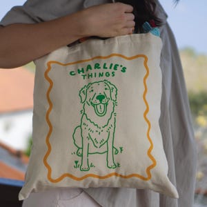 May include: A cream-colored canvas tote bag featuring a green line-art illustration of a dog sitting inside a wavy orange border. The text "CHARLIE'S THINGS" is printed in green above the dog. The bag has two fabric handles and a simple design.