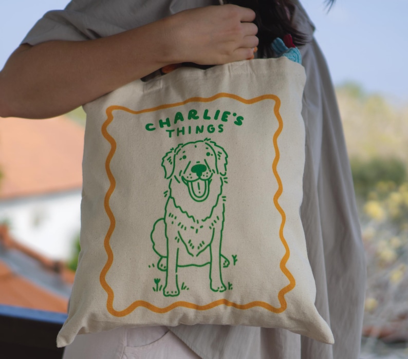 May include: A cream-colored canvas tote bag featuring a green line-art illustration of a dog sitting inside a wavy orange border. The text "CHARLIE'S THINGS" is printed in green above the dog. The bag has two fabric handles and a simple design.
