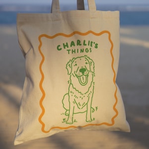 May include: A beige canvas tote bag featuring a green line drawing of a dog sitting down. Above the dog, the text reads CHARLIE'S THINGS in green capital letters. The entire design is enclosed within an orange wavy-lined rectangular border.