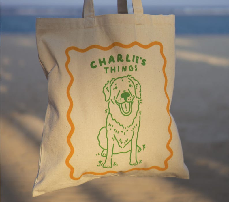 May include: A beige canvas tote bag featuring a green line drawing of a dog sitting down. Above the dog, the text reads CHARLIE'S THINGS in green capital letters. The entire design is enclosed within an orange wavy-lined rectangular border.