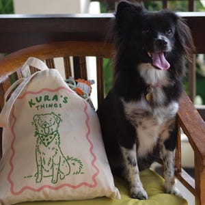 May include: A cream-colored canvas drawstring bag featuring a green line-art illustration of a dog wearing a polka-dot bandana, with the text KURA'S THINGS printed above in green, all framed by a wavy pink border on the front of the fabric bag.