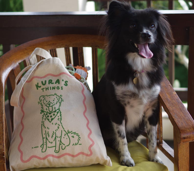 May include: A cream-colored canvas drawstring bag featuring a green line-art illustration of a dog wearing a polka-dot bandana, with the text KURA'S THINGS printed above in green, all framed by a wavy pink border on the front of the fabric bag.