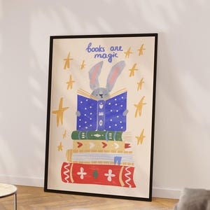 Op de afbeelding: Een ingelijste print met een illustratie. Een grijs konijn leest een blauw gestippeld boek bovenop een stapel kleurrijke boeken. De tekst 'books are magic' staat erboven, met gele steraccenten. Een charmant stuk.